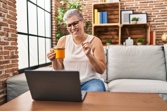 Middle age woman using laptop at home doing video call smiling happy pointing with hand and finger to the side