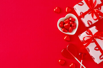 Saint Valentines Day concept. Top view composition made of gift boxes and heart shaped saucers with confectionery chocolate candies and lollipops on red background. Flat lay with copy space.