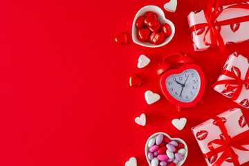 Valentines Day concept. Top view composition made of alarm clock, gift boxes, heart shaped saucers with chocolate candies and marshmallow on red background with copy space. Love Valentines card idea.