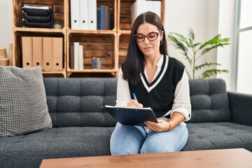 Young beautiful hispanic woman psychologist writing on document at psychology clinic