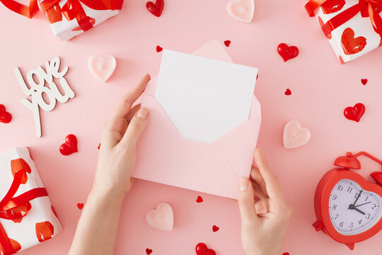 Valentines Day concept. First person top view photo of girl holding envelope with letter over gift boxes, heart shaped candles, red alarm clock, inscription love, baubles on pastel pink background.