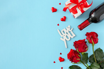 Valentines Day concept. Flat lay composition made of red roses, wine bottle, gift box, red hearts, inscription love you on pastel blue background with copy space. Lovers holiday card idea.