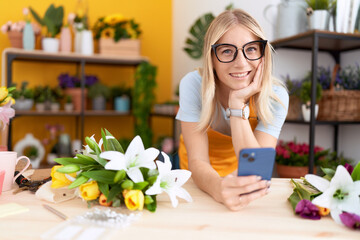 Young blonde woman florist using smartphone leaning on table at flower shop