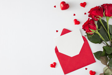 Valentines Day concept. Top view photo of open envelope with white card, red flowers and shaped hearts on white background with copy space. Lovers holiday card idea.