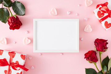 St Valentine's Day concept. Top view composition made of red roses, gift boxes, heart shaped candles on pastel pink background and white frame in the middle. Lovers holiday card idea.