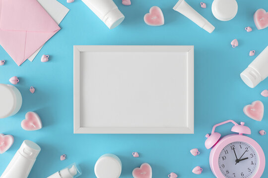 Mother day eve concept. Creative layout made of cosmetic bottles without label, alarm clock, envelope, heart shaped candles on blue background and white frame in the middle. Women skin care idea.
