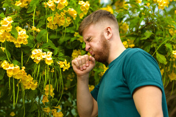 Portrait of handsome young allergic man is suffering from pollen allergy or cold on natural flowers, flowering tree background at spring or sunny summer day, sneezes, blowing his runny nose rubs eyes