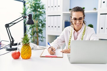 Young hispanic woman wearing nutritionist uniform writing on clipboard at clinic