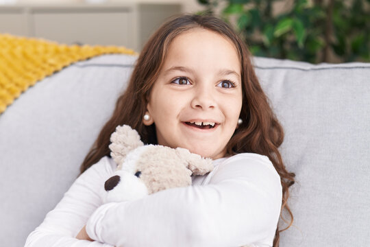 Adorable hispanic girl hugging teddy bear sitting on sofa at home