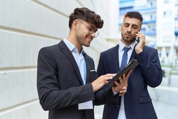 Two hispanic men business workers using touchpad talking on smartphone at street