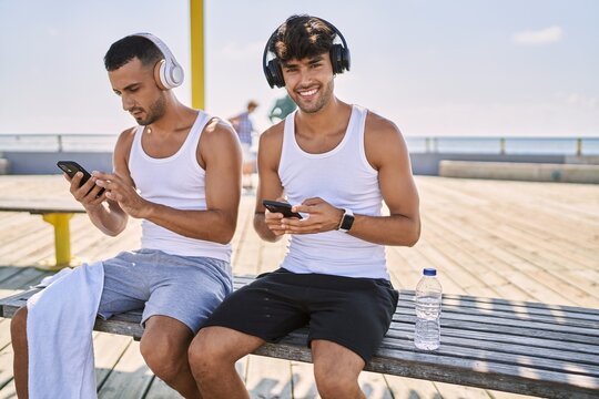 Two hispanic men sporty couple using smartphone and headphones sitting at seaside