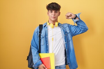 Hispanic teenager wearing student backpack and holding books strong person showing arm muscle,...