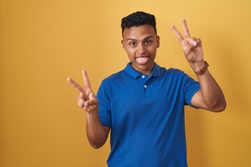 Young hispanic man standing over yellow background smiling with tongue out showing fingers of both...