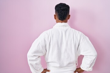 Young hispanic man wearing beauty face mask and bath robe standing backwards looking away with arms on body
