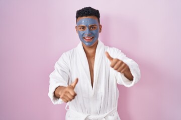 Young hispanic man wearing beauty face mask and bath robe approving doing positive gesture with...