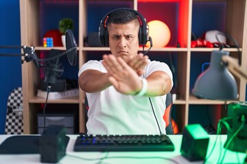 Young hispanic man playing video games rejection expression crossing arms and palms doing negative sign, angry face