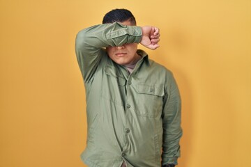 Hispanic young man standing over yellow background covering eyes with arm, looking serious and sad....