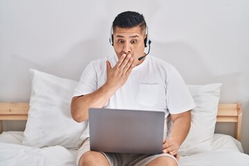 Hispanic young man using computer laptop on the bed covering mouth with hand, shocked and afraid for mistake. surprised expression