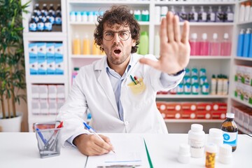Hispanic young man working at pharmacy drugstore doing stop gesture with hands palms, angry and...