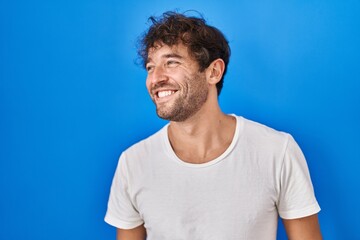 Hispanic young man standing over blue background looking away to side with smile on face, natural...