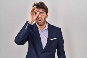 Hispanic business man wearing glasses doing ok gesture shocked with surprised face, eye looking through fingers. unbelieving expression.
