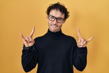 Hispanic man standing over yellow background shouting with crazy expression doing rock symbol with hands up. music star. heavy music concept.