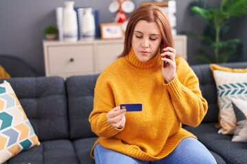 Young beautiful plus size woman talking on smartphone holding credit card with serious expression at home