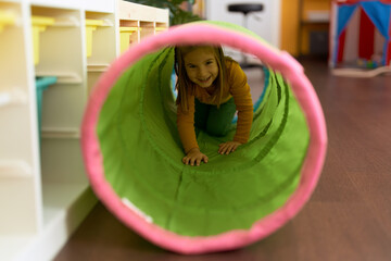 Adorable hispanic girl crawling inside tunnel toy at kindergarten