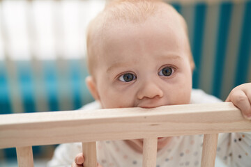 Adorable caucasian baby standing on cradle biting wooden balustrade at bedroom