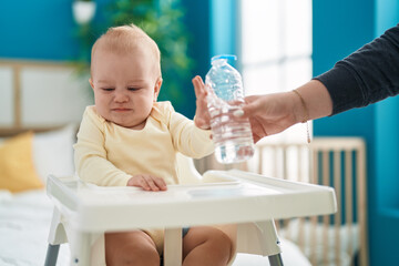 Adorable caucasian baby sitting on highchair rejecting bottle of water at bedroom