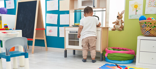 Adorable hispanic toddler playing with play kitchen standing at kindergarten