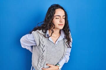 Fototapeta premium Young brunette woman standing over blue background suffering of backache, touching back with hand, muscular pain