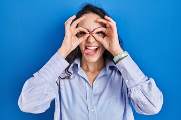 Young brunette woman standing over blue background doing ok gesture like binoculars sticking tongue out, eyes looking through fingers. crazy expression.