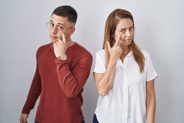 Mother and son standing together over isolated background pointing to the eye watching you gesture, suspicious expression