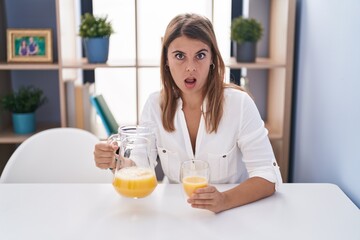 Young hispanic woman drinking glass of orange juice in shock face, looking skeptical and sarcastic, surprised with open mouth