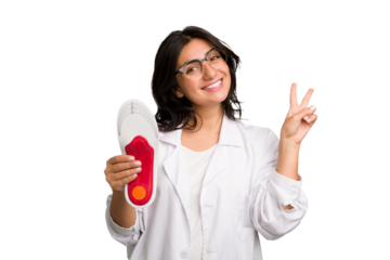 Young indian chiropodist woman holding a insoles for shoes cut out isolated joyful and carefree showing a peace symbol with fingers.