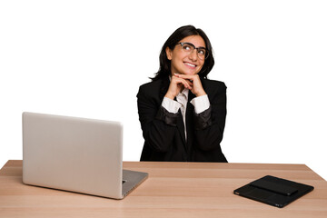 Young indian woman in a table with a laptop and tablet isolated keeps hands under chin, is looking happily aside.