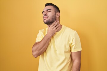 Young hispanic man standing over yellow background touching painful neck, sore throat for flu, clod...