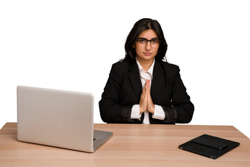Young indian woman in a table with a laptop and tablet isolated praying, showing devotion,...