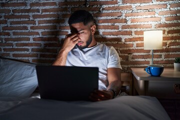 Young latin man tired using laptop sitting on bed at bedroom