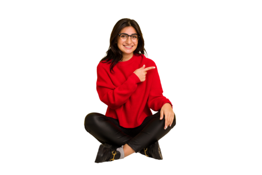Young indian woman sitting on the floor cut out isolated smiling and pointing aside, showing something at blank space.