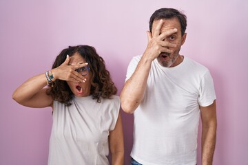Middle age hispanic couple together over pink background peeking in shock covering face and eyes with hand, looking through fingers with embarrassed expression.