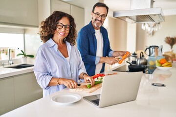 Middle age hispanic couple cooking and using laptop at kitchen