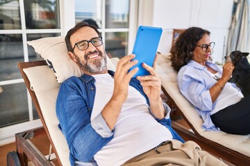 Middle age hispanic couple using touchpad lying on hammock with dog at terrace