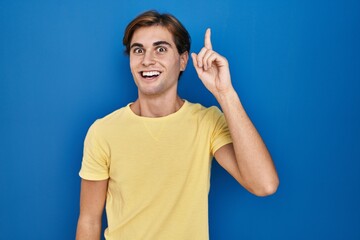 Young man standing over blue background pointing finger up with successful idea. exited and happy. number one.