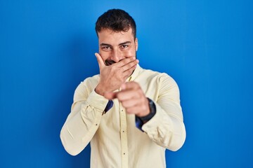 Handsome hispanic man standing over blue background laughing at you, pointing finger to the camera...