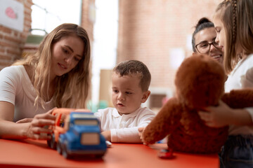 Teachers with boy and girl playing with cars toy and doll sitting on table at kindergarten