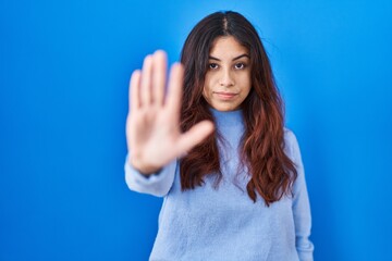 Hispanic young woman standing over blue background doing stop sing with palm of the hand. warning...