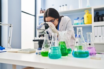 Young hispanic woman wearing scientist uniform using microscope at laboratory