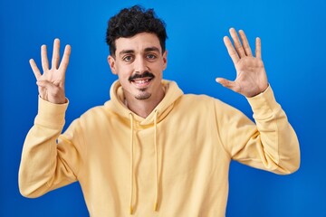 Hispanic man standing over blue background showing and pointing up with fingers number nine while...